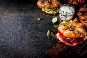 Variety of homemade bagels sandwiches with sesame and poppy seeds, cream cheese,  ham, radish, arugula, cherry tomatoes, cucumbers, with ingredients on dark concrete background copy space