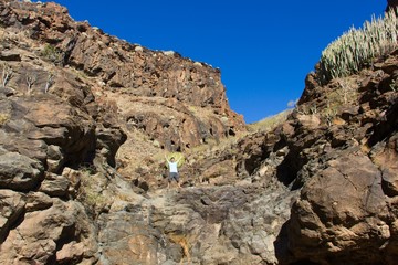Young man with arms up celebrates from top of rock in the middle of arid mountain landscape in Canary Islands, Spain