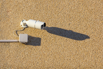 A white street videocamera with cable and a box hangs on a concrete wall and casts a shadow over...
