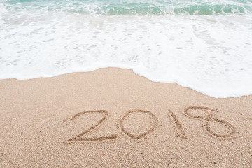 Happy new year 2018 concept. Number 2018 written on sandy beach with blue sea waves and foam.