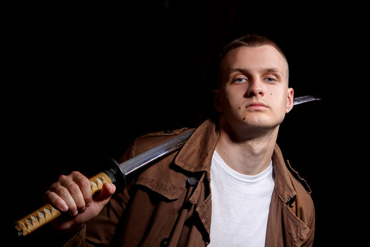 Young man dressed in a brown coat and white t-shiert, holding a katana.Modern samuria.Fashion portrait.Urban warior.Dramatic light