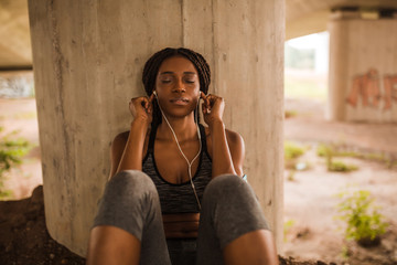 Young black woman in sportswear listening to her favorite music while sitting next to pillar below a bridge