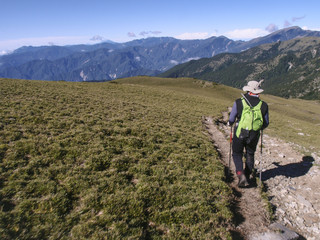 Obraz premium A male hiker in a hat holding trekking poles and walking on a trail. Background is the beautiful alpine landscape of Taiwan with a clear blue sky.