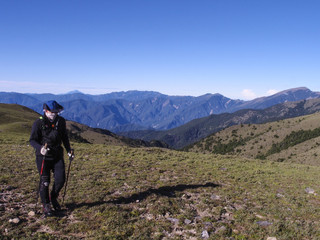 Fototapeta na wymiar A male hiker in a hat holding trekking poles and walking on a trail. Background is the beautiful alpine landscape of Taiwan with a clear blue sky.