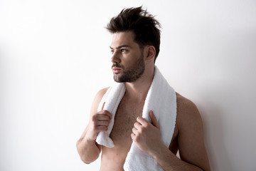 Young sleepy bearded man with tousled hair is standing against light wall and holding white towel over his neck. He is looking aside thoughtfully. His torso is naked