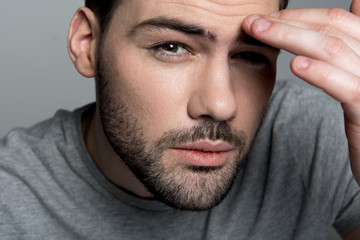 Obraz premium Ache concept. Close up portrait of young exhausted man is looking at camera while suffering from headache. He is touching his forehead. Focus on his face