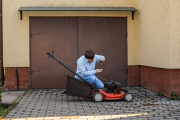 Young man in jeans and shirt checks the oil level in the lawn mower opposite the garage in the summer.