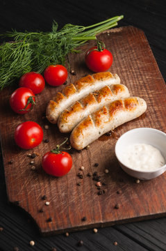 Grilled sausages grilled on a brown wooden board on a wooden background, six cherry tomatoes, greens, parsley dill, pepper, sauce. Place under the text and logo. Top view. Flat lay.