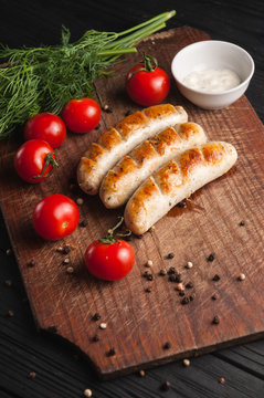 Grilled sausages grilled on a brown wooden board on a wooden background, six cherry tomatoes, greens, parsley dill, pepper, sauce. Place under the text and logo. Top view. Flat lay.