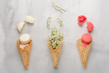 Flat-lay of a sweet cone of wafers with white flowers and macaroons on a light background, top view. The concept of spring or summer mood