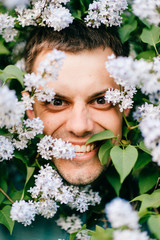 Obraz premium Closeup odd unusual male portrait outdoor at nature in beautiful lilac bushes. Adult funny man face grimace surrounded by blooming flowers. Emotions and facial expxression. Clowning and fooling.