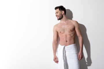 Full of joy. Young happy topless guy with stubble is standing with towel on hips and looking aside with wide smile. Copy space in the left side. Beauty concept © Yakobchuk Olena