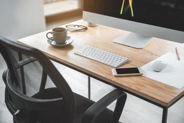 Close up of modern mobile locating near keyboard of computer and cup of coffee on table. Cozy chair is near it. Office appliance concept