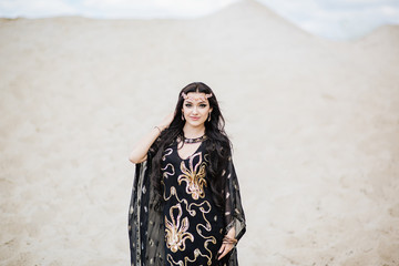 Dancer bellydance and Khaleegy (Khaliji) in a black suit in the desert against the background of sand. Image of Persian Gulf. Beautiful nature. Belly dance. A girl with an exotic look. Eastern beauty.