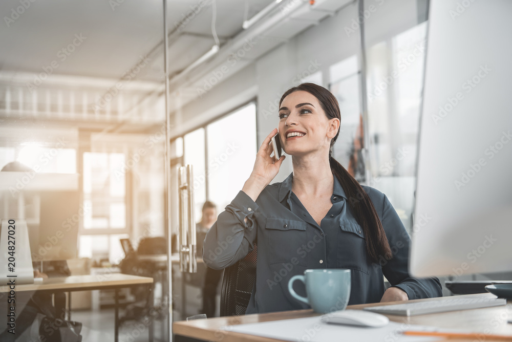 Poster portrait of beaming businesswoman talking by phone while working with computer at table