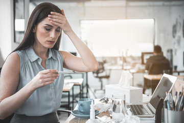 Portrait of serene ill woman looking at clinical thermometer while holding forehead by hand. Flue during job concept