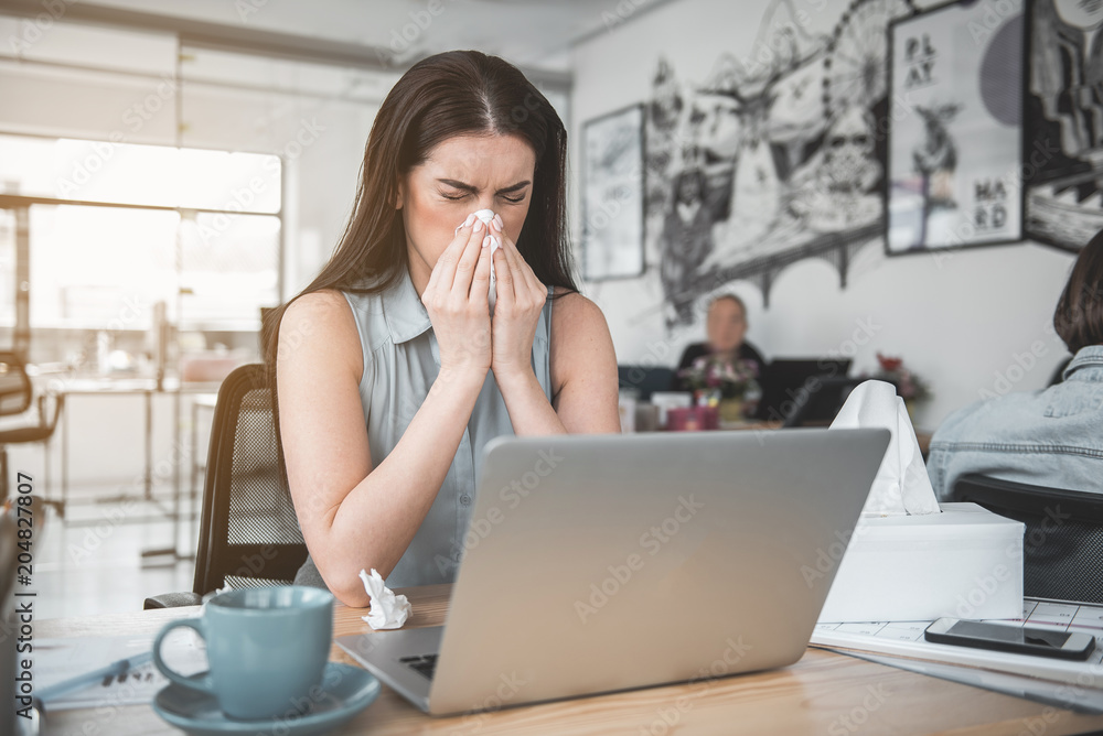 Poster portrait of disappointed businesswoman blowing nose into handkerchief while using notebook computer.