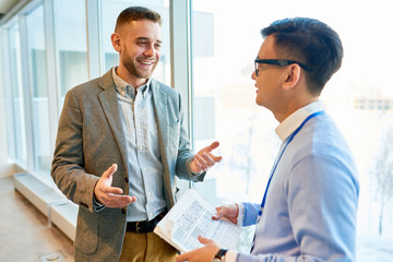 Waist up portrait of successful modern businessman talking to Asian colleague and smiling happily