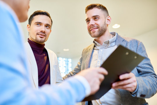 Low Angle Portrait Of Three Handsome Business People Holding Clipboard And Smiling While Discussing Financial Strategies In Modern Office Building