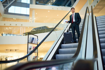 Full length wide angle portrait of successful Asian businessman descending on escalator holding...