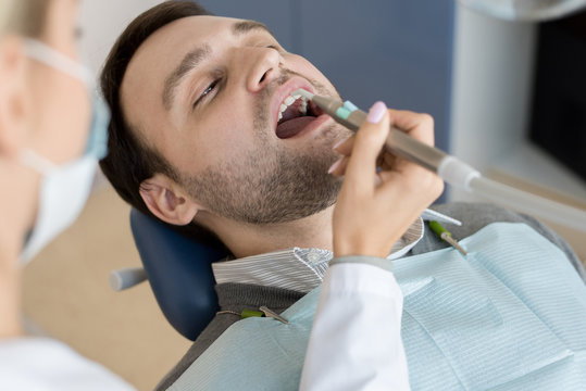 Portrait of mature man sitting in dental chair with mouth open while female dentist treating teeth in modern clinic