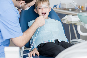 Portrait of dentist treating teeth of cute little boy sitting in dental chair, filling cavities and doing professional cleaning