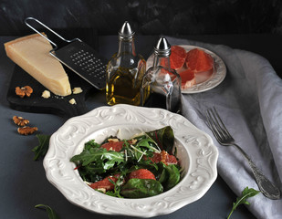 Salad with pink grapefruit. Near the board with a piece of Parmesan cheese and a plate with slices of grapefruit. In the frame walnuts, bottles with olive oil, balsamic, fork and napkin. Close-up.
