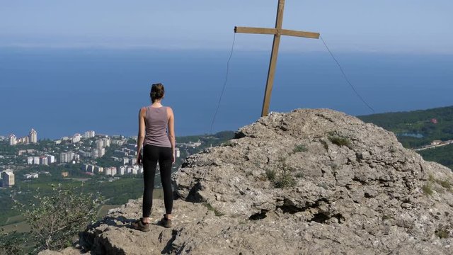 The view from the back to the girl standing on a rocky mountain top with the cross and looking into the distance. Yalta, Crimea