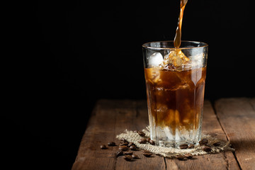 Ice coffee in a tall glass over and coffee beans on a old rustic wooden table. Cold summer drink on a dark background with copy space. The process of pouring drink from a coffee pot into a glass