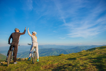 happy couple goes on a mountain road in the woods on bikes with helmets giving each other a high...