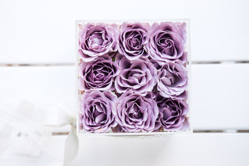 Pastel purple hued roses in clear acrylic crystal flower box. Square glass gift box. On white wooden table from above, copyspace