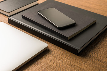 close up view of laptop, pile of black notebooks and smartphone on wooden tabletop