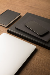 close up view of laptop, pile of black notebooks and smartphone on wooden tabletop