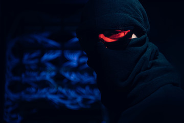 obscured view of hacker in eyeglasses and mask looking at camera with cables on background