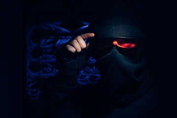 obscured view of hacker in eyeglasses and mask looking at camera with cables on background