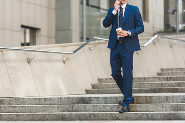 cropped shot of businessman in stylish suit with coffee to go talking by phone while walking by...