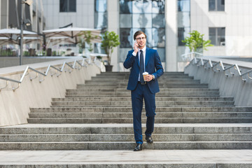 smiling young businessman in stylish suit with coffee to go talking by phone while walking by...