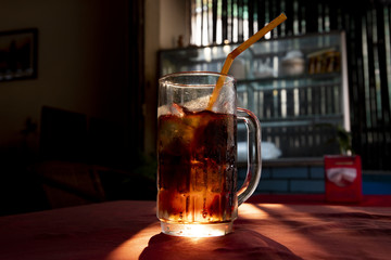 Coke inglass with straw. Brown sparkling beverage with sun backlight. Sparkling drink with ice in glass
