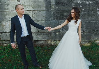 Fototapeta na wymiar Wedding couple is walking outdoors in sunny green summer day. Medieval stone walls with old renaissance palace on background. Bride is satin lace dress is holding groom hand. Old castle architecture.