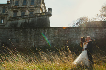 Obraz premium Wedding couple is walking outdoors in sunny day. Medieval stone walls with old renaissance palace on background. Bride is satin lace dress is embracing groom. Old architecture and dry high grass.