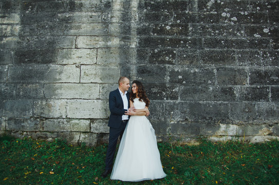 Wedding couple is hugging outdoors in sunny green summer day. Medieval stone walls with old renaissance palace on background. Bride is satin lace dress is holding groom hand. Old castle architecture.
