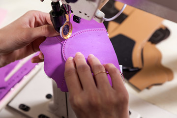 Close up a young woman working as a seamstress in a purple unifrome sews genuine leather children's shoes on a sewing machine on a table in a sewing workshop