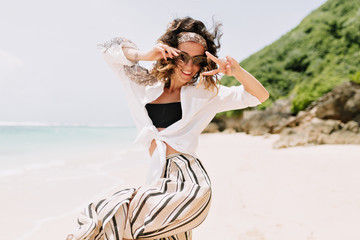 Fototapeta na wymiar Pretty lovely young woman with dark hair with hair ornament wers black sunglasses, white shirt and striped pants jumping on the white beach with green rocks near the ocean in sunlight