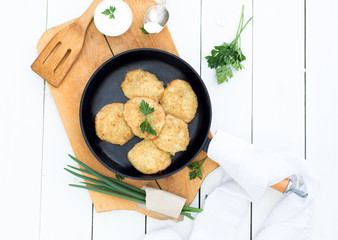 Potato pancakes in frying pan with sour cream and greens on white wooden table.