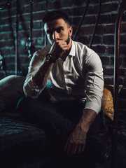 A handsome man with a beard and a white shirt sits on the bed and looks thoughtfully at the camera. Intimate atmosphere and twilight. - 204864843