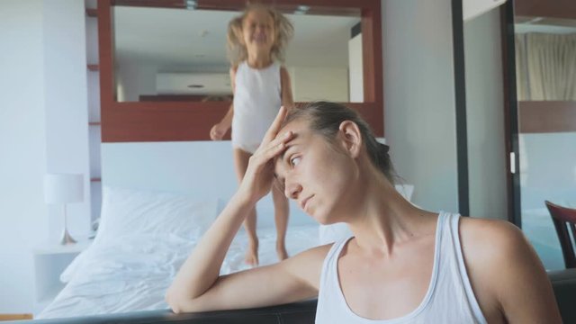 Upset mother with hyperactive child jumping on bed on the background