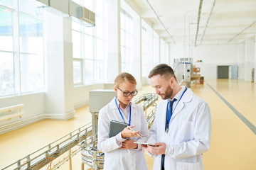 Fototapeta premium Confident inspectors wearing white coats using digital tablet while standing at spacious packaging department of milk factory