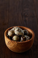 Fresh quail eggs in a wooden bowl on a homespun napkin over dark wooden background, top view, close-up