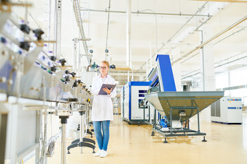 Concentrated fair-haired worker wearing white coat standing at conveyor belt and controlling production process at spacious dairy factory