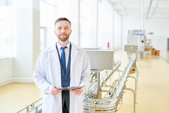 Portrait shot of handsome bearded inspector wearing suit and white coat looking at camera while carrying out inspection at spacious dairy plant